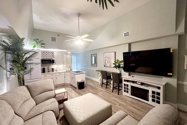 Open-plan living room with beige seating, kitchen with white cabinets, wall-mounted TV, and ceiling fan