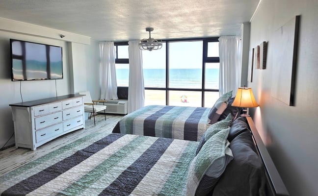 Beachfront hotel room with two beds, ocean view through large windows, white dresser, and TV.
